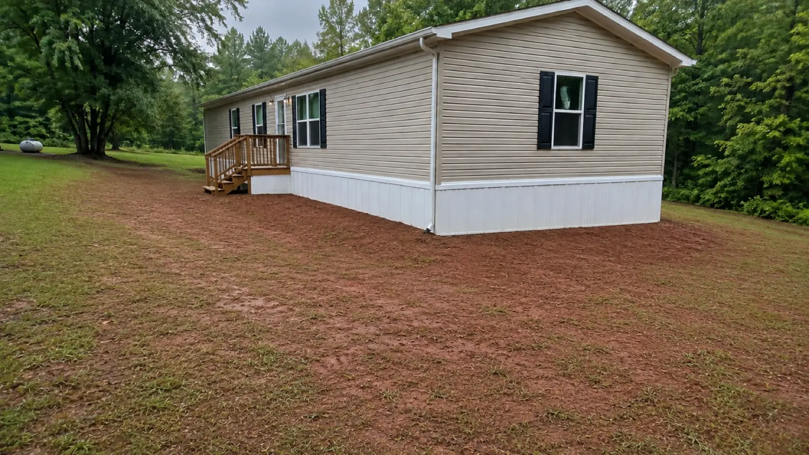 A newly set manufactured home on a graded red-clay pad that slopes away from the skirting, seeded and strawed, dry after rain.