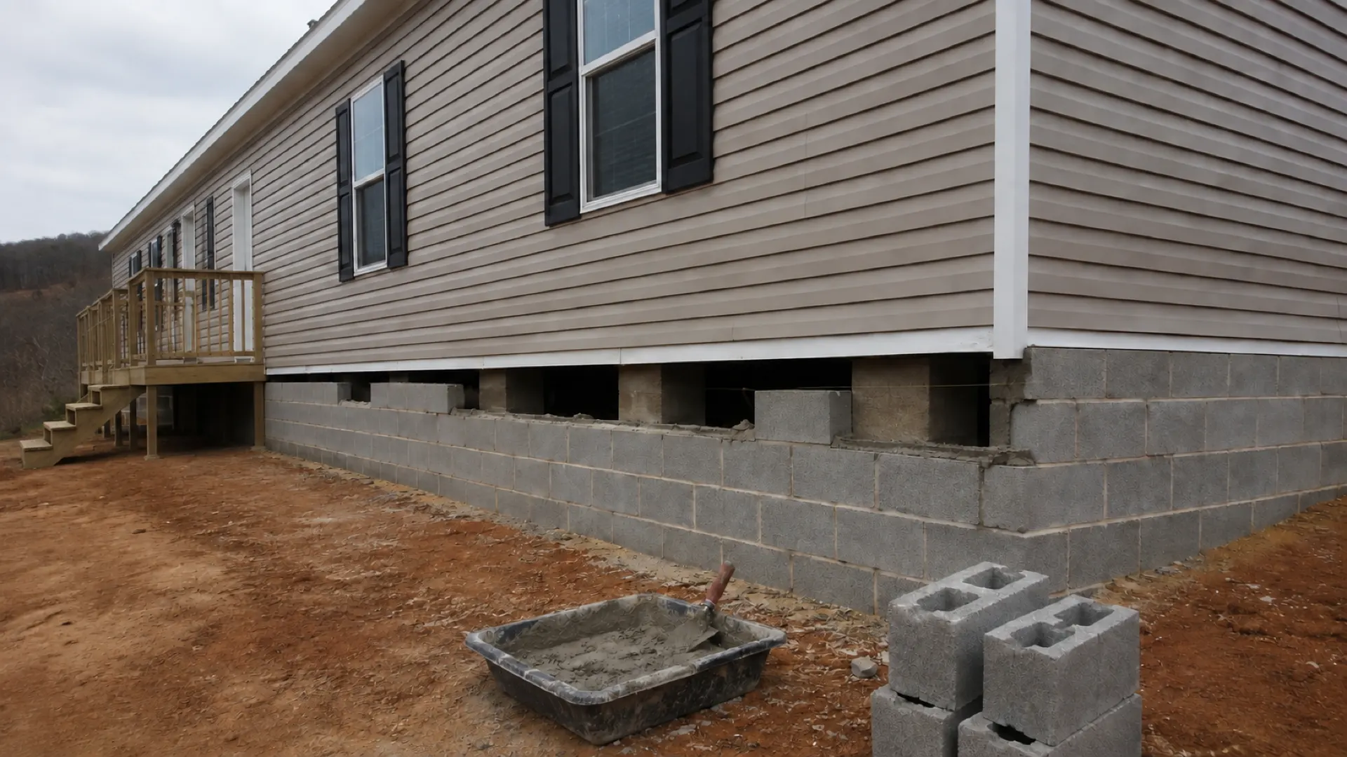 A block curtain wall being laid in mortar under the edge of a set doublewide, with blocks and a mortar tub on the ground.