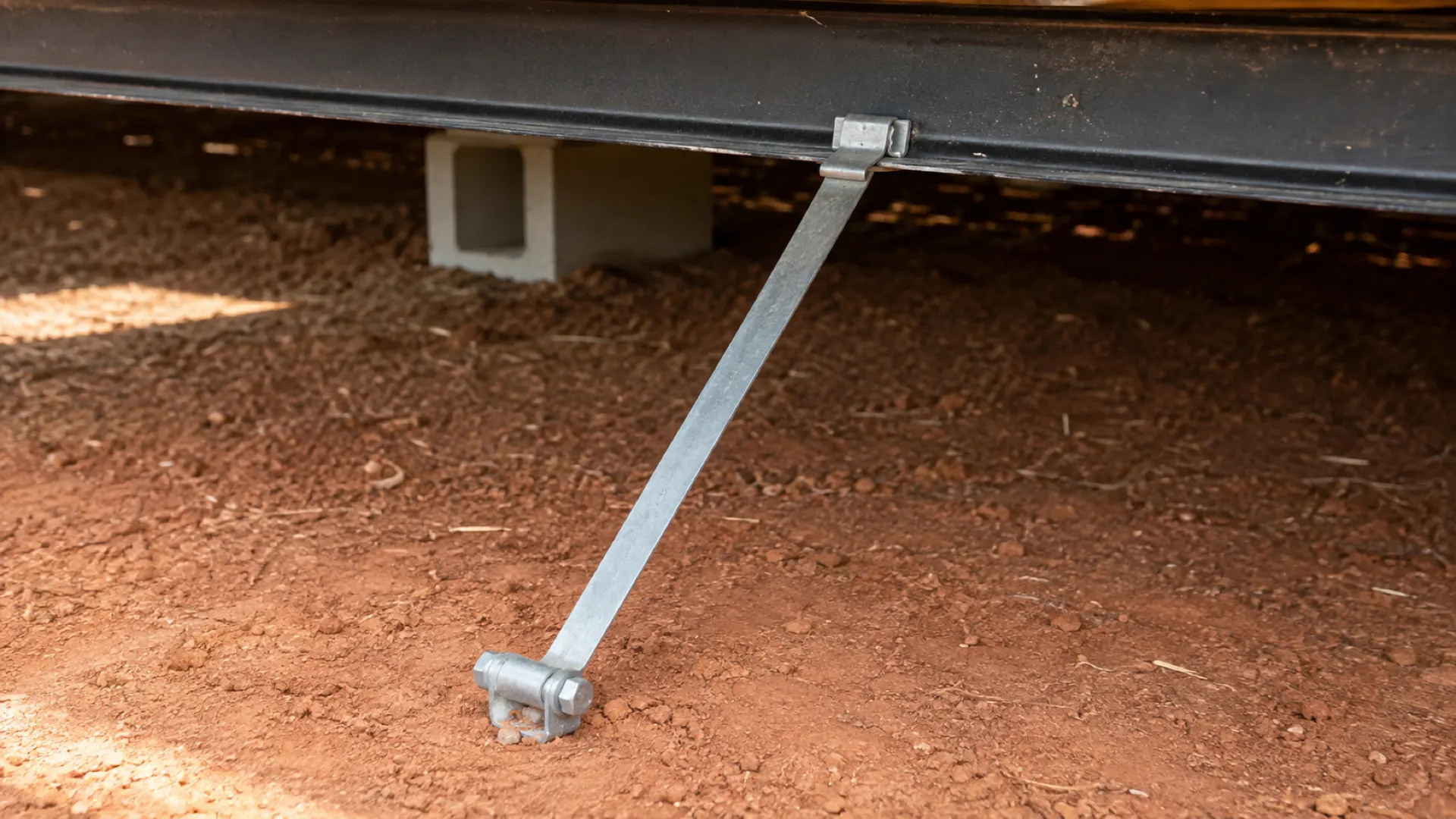 A galvanized tie-down strap running straight and tight from a manufactured home frame to a ground anchor set flush with the soil.